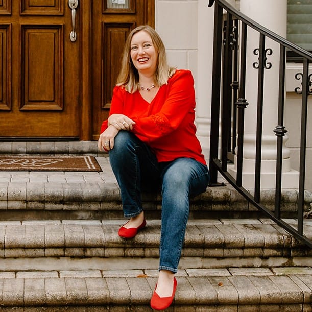 Dani sitting on the steps of a home, wearing a red blouse and jeans, smiling at the camera.
