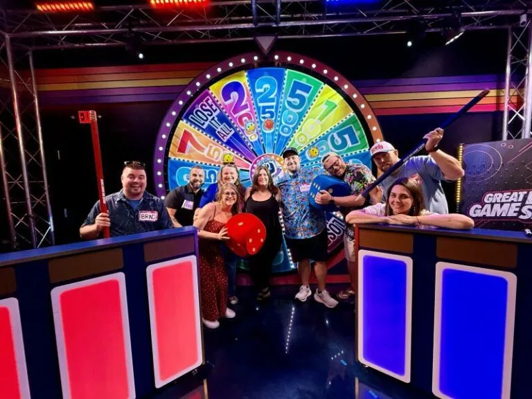 A group of nine adults posing together on a colorful game show set. They are standing in front of a large, brightly lit spinning prize wheel divided into multicolored sections with numbers and labels. Several of the adults hold oversized props, including giant red and blue dice and a long red pole. There are wo podiums with glowing red and blue panels frame the group on either side. The set is decorated with stage lighting and a “Great Games” sign in the background, creating a lively, playful atmosphere.