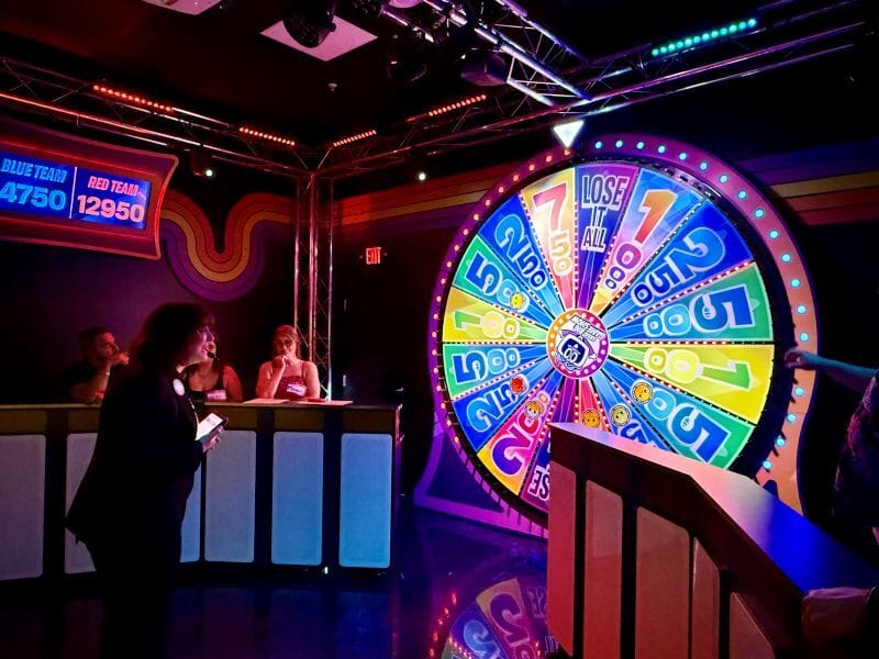 A large colorful prize wheel with multicolored sections. To the left, two teams stand behind podiums under bright stage lighting. A scoreboard above them shows the red team leading the blue team. A host stands in the middle holding a tablet, facing the wheel as the game continues.